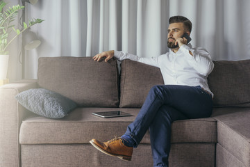 Young bearded businessman, dressed in white shirt and blue pants, sitting in room on brown sofa and...