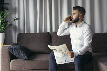 Young bearded businessman, dressed in a white shirt, sitting in a room on a brown sofa and talking on a cell phone while holding a newspaper. In the background gray curtains. Nearby is a house plant.