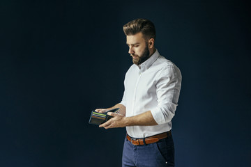 Young bearded businessman, dressed in a white shirt and dark blue trousers, standing and holding an open wallet with a credit card. In the background dark blue wall. A man looks into an empty wallet.