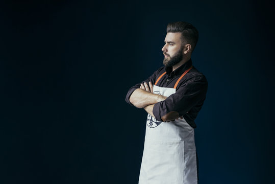 Side view. A young bearded man, dressed in a dark brown shirt and bright apron, standing with his arms folder. On background dark blue wall.