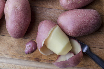Partly peeled potato with intact ones with peeling knife on wooden cutting board from above