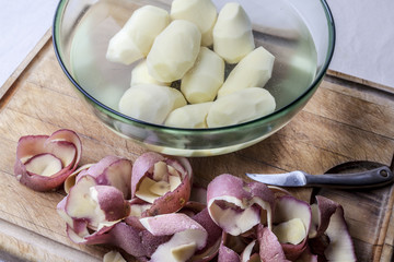 Peeled potatoes in glass bowl with water peels aside and peeling knife on wooden cutting board from high angle