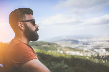 Fototapeta na wymiar Summer sunny day. Side view of a young bearded man in sunglasses and a brown T-shirt standing on the highest point, looking at the landscape from a bird's flight. Tourist enjoying the view.
