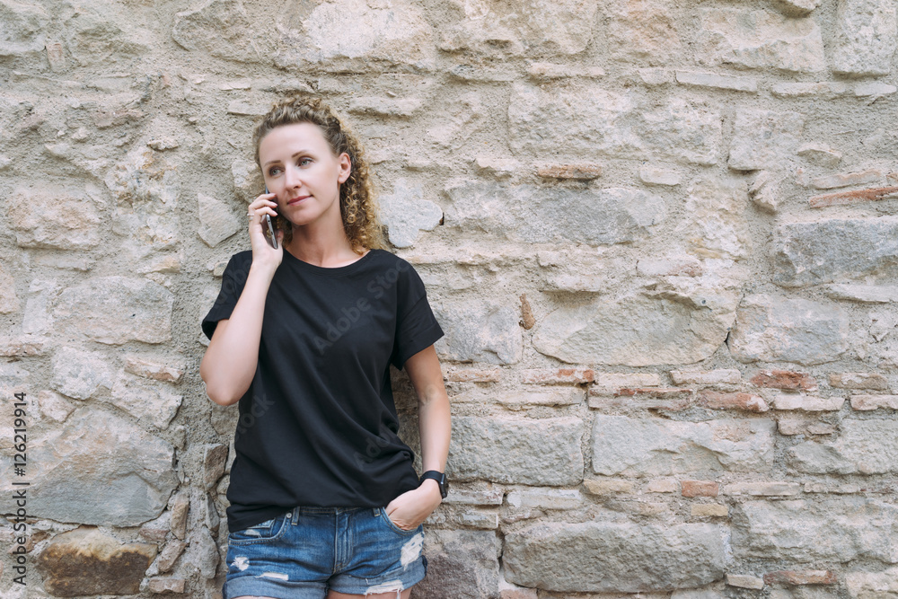 Wall mural Front view of a young woman dressed in a black T-shirt and denim shorts, standing and talking on the phone. In the background is bright stone wall. Girl uses the gadget.