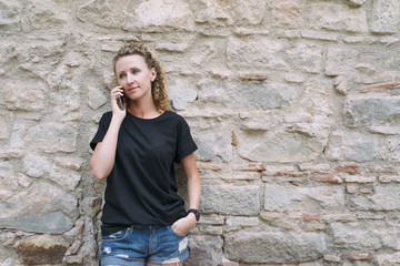 Front view of a young woman dressed in a black T-shirt and denim shorts, standing and talking on the phone. In the background is bright stone wall. Girl uses the gadget.