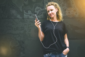 Front view of young woman with curly hair,dressed in black T-shirt and jeans,standing against background of black walls with stone texture and listening to music on headphones while holding smartphone