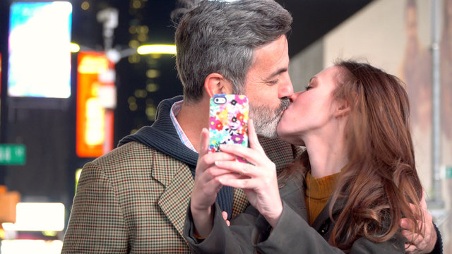 Young attractive couple on date night in Times Square New York City, Manhattan - taking selfie photo kissing with smart cell phone to share on social media for friends to tag and like.