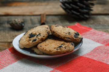 Chocolate cookies in a white plate on a checkered tablecloth on wooden background. Near cones and cinnamon stick, roughly. The theme of the new year holiday