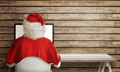 Santa Claus work on computer at his home. Wooden wall and table with free space for text.
