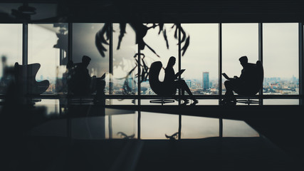 Silhouettes of two business people man and woman working on their gadgets sitting on armchairs in office interior with a lot of reflections, with huge windows and winter cityscape outside