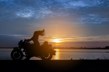 Silhouette biker woman with his motorbike beside the natural lake with sunset background.