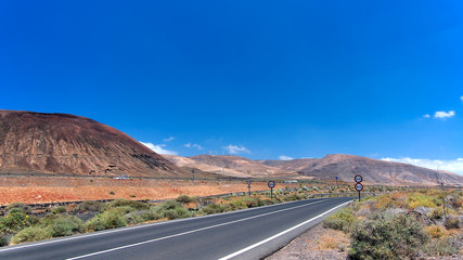 Speed limit signs on asphalt road disappearing over the horizon through volcano mountain hillsides. White clouds on a blue sky. Lanzarote, Canary Islands, Spain