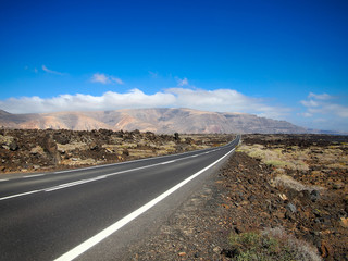 Asphalt road disappearing over the horizon through volcano mountain hillsides. White clouds on a blue sky. Lanzarote, Canary Islands, Spain