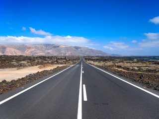 Fototapeta na wymiar Asphalt road disappearing over the horizon through volcano mountain hillsides. White clouds on a blue sky. Lanzarote, Canary Islands, Spain
