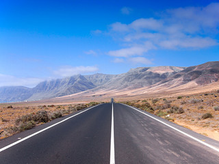 Asphalt road disappearing over the horizon through volcano mountain hillsides. White clouds on a blue sky. Lanzarote, Canary Islands, Spain