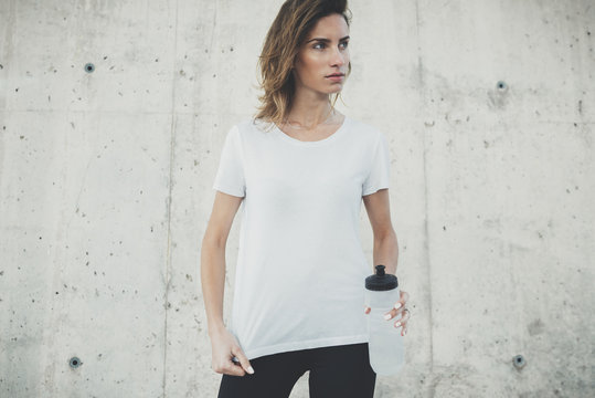 Young fitness girl wearing blank white t-shirt with empty area for design or content, mockup of template white t-shirt, female jogger resting after active workout outside, concrete wall in background