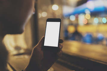 Young hipster man sitting at cafe with cellphone, mockup of template mobile, closeup of male hand holding and using modern smartphone with blank screen for text or content, with night light bokeh