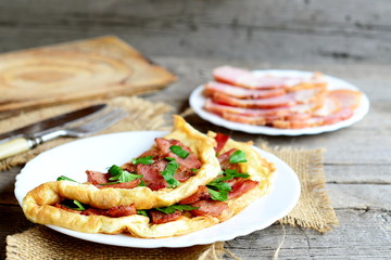 Homemade bacon omelette recipe. Beautiful omelette stuffed with fried bacon and fresh parsley on a plate, bacon slices, fork, knife, cutting board, on old wooden background. Delicious breakfast recipe