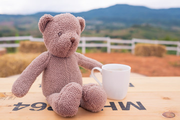 Bear doll with a cup of coffee on wooden table against mountain background. Vintage color.