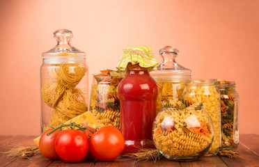 Various shapes pasta in jars, bottle ketchup, tomato on brown.