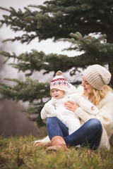 Fototapeta premium Beautiful smiling young woman with adorable baby boy sitting on the grass near a nice fir tree. Mother with child walking in the park on a cold day. Motherhood and lifestyle concept