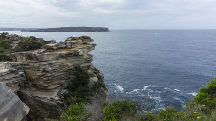 Fototapeta na wymiar Steep rocky coast of Australia and the blue sea waves breaking on the rocks, the entrance to Sydney Harbour, Sydney