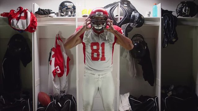 A football player sitting in his locker puts on his helmet and gets ready to play