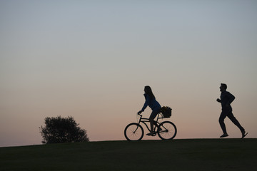 Silhouette of a couple walking through a park in San Diego at dusk. The woman is riding a bike and the man is walking. 