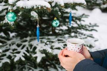 man holding a cup of hot tea in his hands next to the Christmas tree in winter forest in the New Year holidays