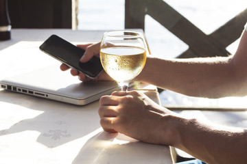 Cropped image of man's hand with telephone and glass of white wine near the sea.