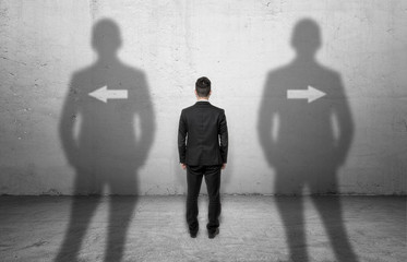 Fototapeta premium Businessman standing in front of a concrete wall with arrows pointing at different directions.