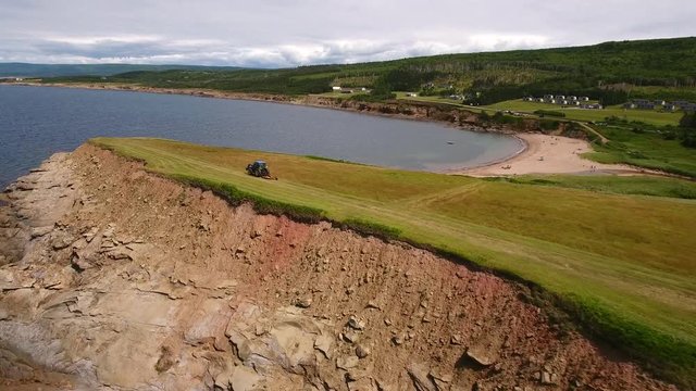 Aerial shot as farmer cuts grass on field by ocean cove