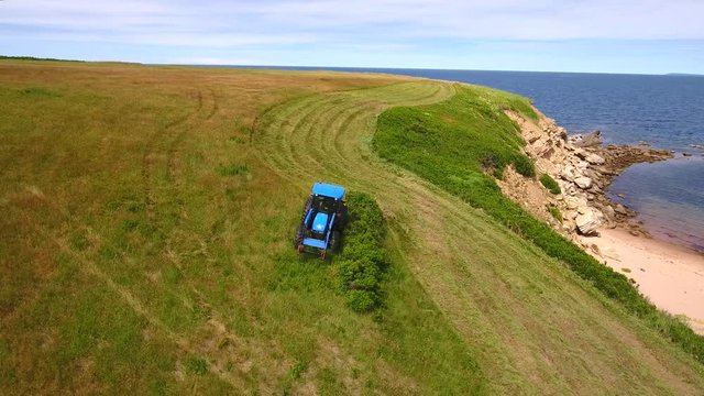 Aerial rotating shot as a farmer cuts grass on a field by ocean cove