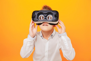 Small boy in white shirt looking at camera through virtual reality headset isolated on orange background.