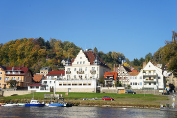 Obraz premium Kurort an der Elbe/Idyllischer Kurort an der Elbe in der Sächsischen Schweiz in Deutschland, schöne Häuser und bunte Laubwälder, im Vordergrund die Elbe, 