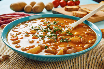 Bean soup in blue dish and wooden spoon surrounded by potatoes, tomatoes, beans in pod, croutons on blue wooden table