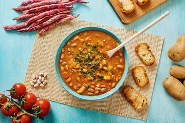 Bean soup in blue dish and wooden spoon, surrounded by potatoes, tomatoes, beans in pod, croutons on blue wooden table