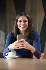 Smiling young woman is using her phone while having a coffee break in a restaurant