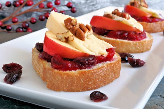 Crostini appetizers with cranberry sauce, apples, brie and pecans on white plate