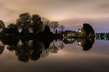 Obraz premium Night view over the Amstel river at Kalfjeslaan, Amsterdam. Still water reflects mirror images of a house and treeline at the East bank (Ouderkerkerdijk). A ferry service (De Smient) runs in summer.