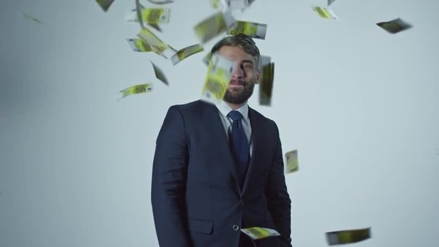 Portrait of satisfied bearded businessman in suit and tie standing isolated on grey background and smiling as money falling on him in slow motion