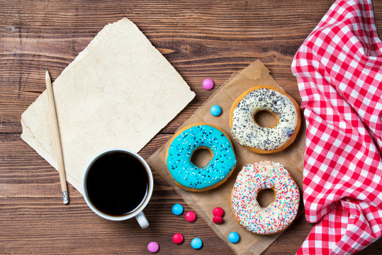 Colorful donuts, cup of coffee, smarties and old blank paper sheet with pencil on wooden table, top view