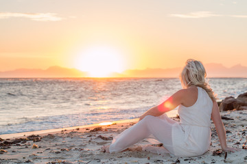 Blonde model sitting on beach looking at sunset.