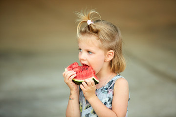 Adorable blonde girl eats a slice of watermelon outdoors.