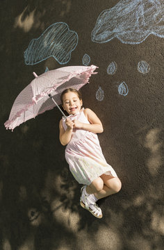 Little girl laying down on asphalt with umbrella after drawing clouds with rain drops with chalk. Concept of child imagination. 