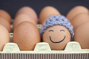 Smiling egg wearing a knitted hat souronded by blank brown eggs.