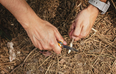 Close-up of male hiker hands sharpen stick with knife on the ground. Concept of travel, extreme and survival. 