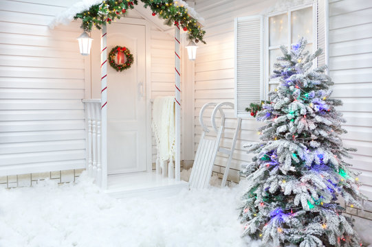 Winter exterior of a country house with Christmas decorations in the American style. Snow-covered courtyard with a porch, tree and wooden vintage sleds.