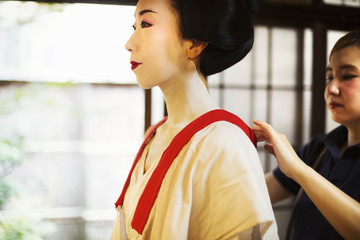 A modern geisha or maiko woman being dressed in traditional fashion in a white shift with loose collar, with white face makeup.