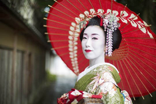 A woman dressed in the traditional geisha style, wearing a kimono and obi, with an elaborate hairstyle and floral hair clips, with white face makeup with bright red lips and dark eyes holding a red paper parasol. 